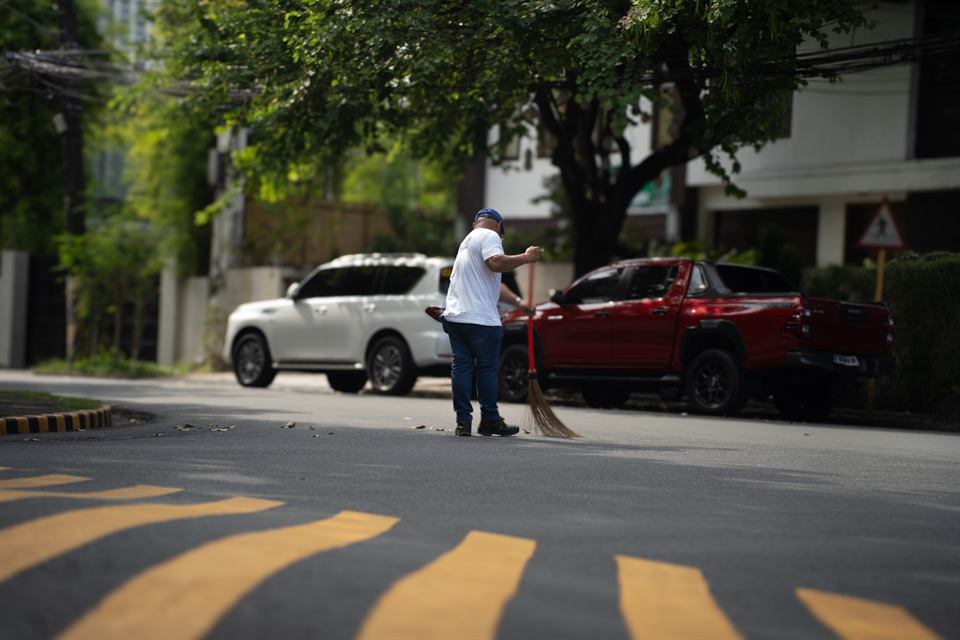 A man sweeping a quiet village street, cars parked under the trees