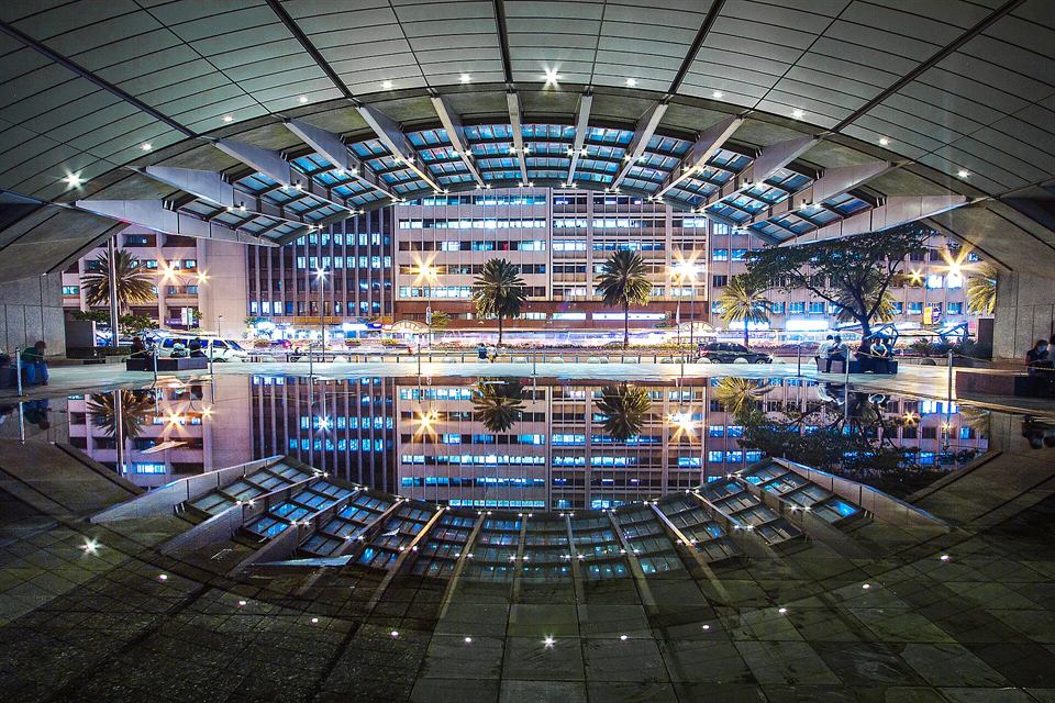 A covered plaza at night, its lit ceiling reflected in a pool, office towers beyond